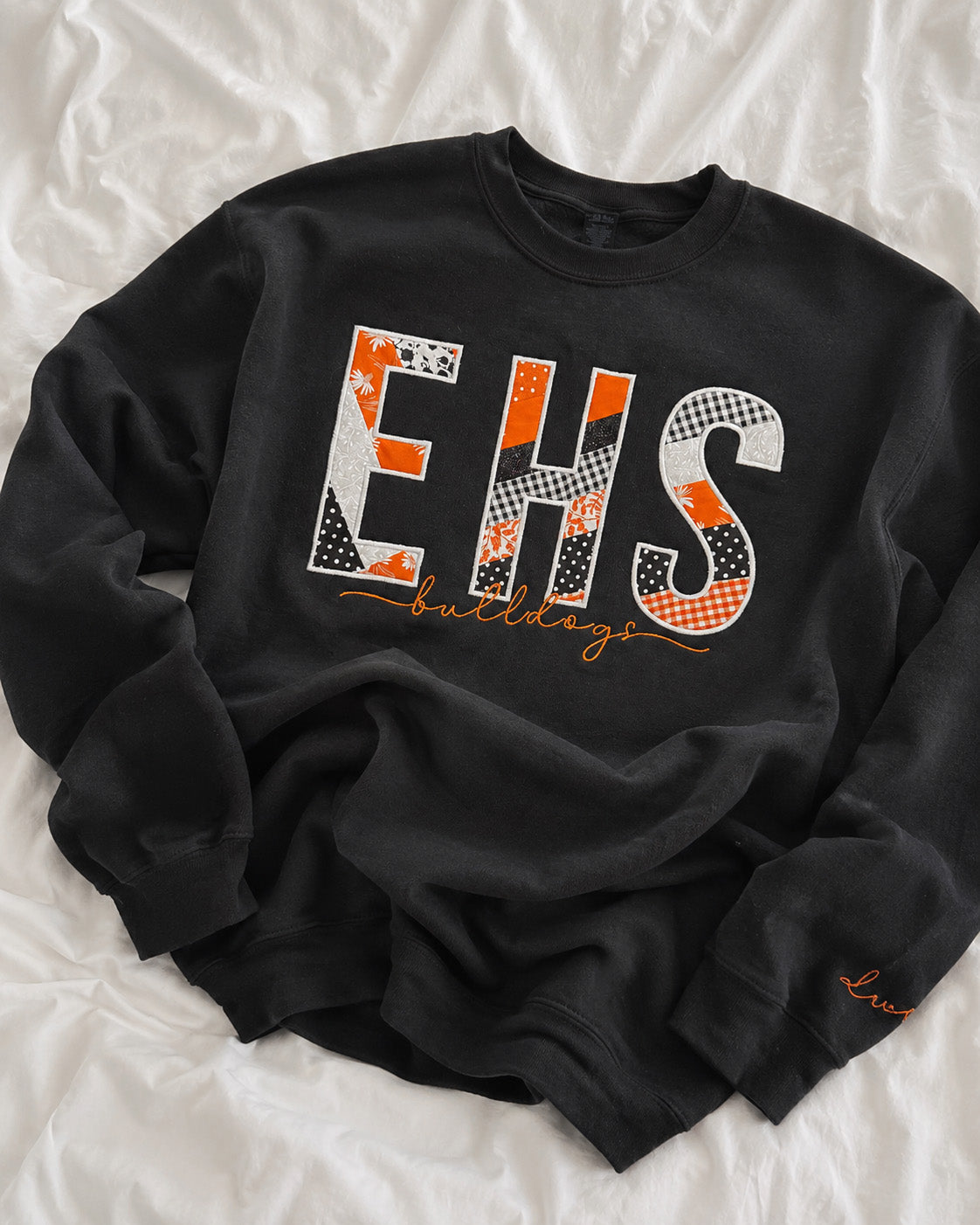 A black crewneck sweatshirt with a large 'EHS' graphic on the chest, laid flat on a white wrinkled fabric background.