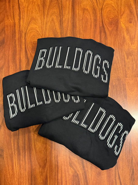 Three black sweatshirts with 'BULLDOGS' printed in white outlined block letters, stacked on a wooden floor.