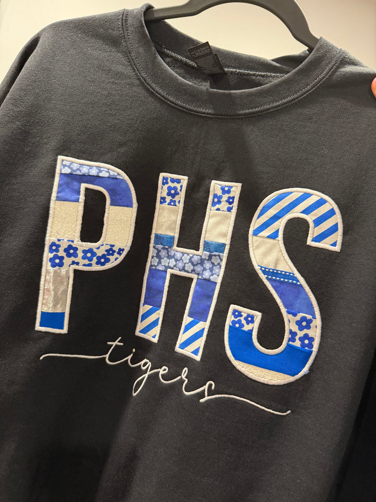A black crewneck sweatshirt hanging on a black hanger, featuring a large patchwork-style 'PHS' graphic in blue, white, and beige, with 'tigers' written in white script underneath.