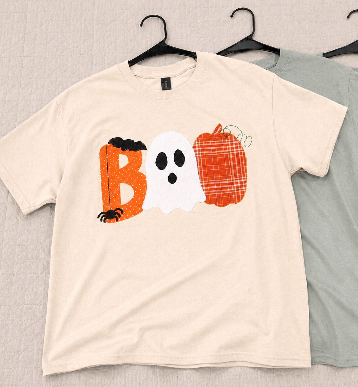 A cream-colored short-sleeve t-shirt hanging on a black hanger, featuring a Halloween graphic that spells 'BOO' with an orange pumpkin, a white ghost, and a plaid pumpkin. A second sage green t-shirt is partially visible behind it on a black hanger against a light gray textured background.