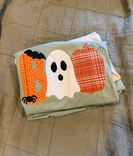 A folded sage green t-shirt featuring a Halloween applique design spelling 'BOO' with a white ghost, orange polka-dot B, and red plaid pumpkin, displayed on a beige quilted blanket.