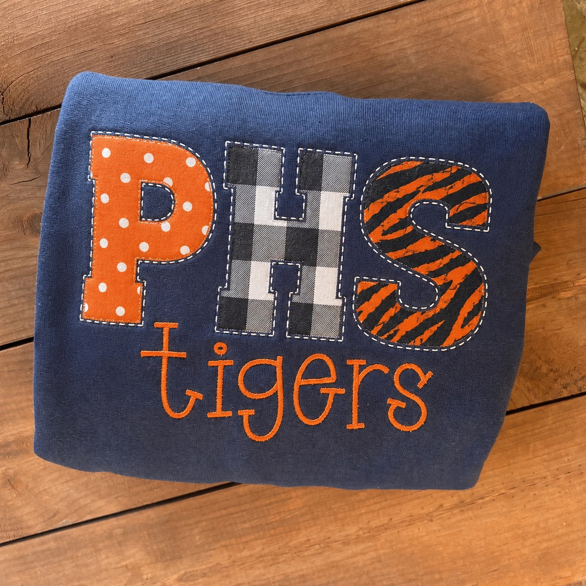 A folded navy blue fabric item, likely a sweatshirt or blanket, featuring a custom applique design that reads 'PHS Tigers'. The letters 'P' and 'S' are filled with orange patterns (polka dots and tiger stripes), while 'H' is in a gray and white buffalo plaid pattern.