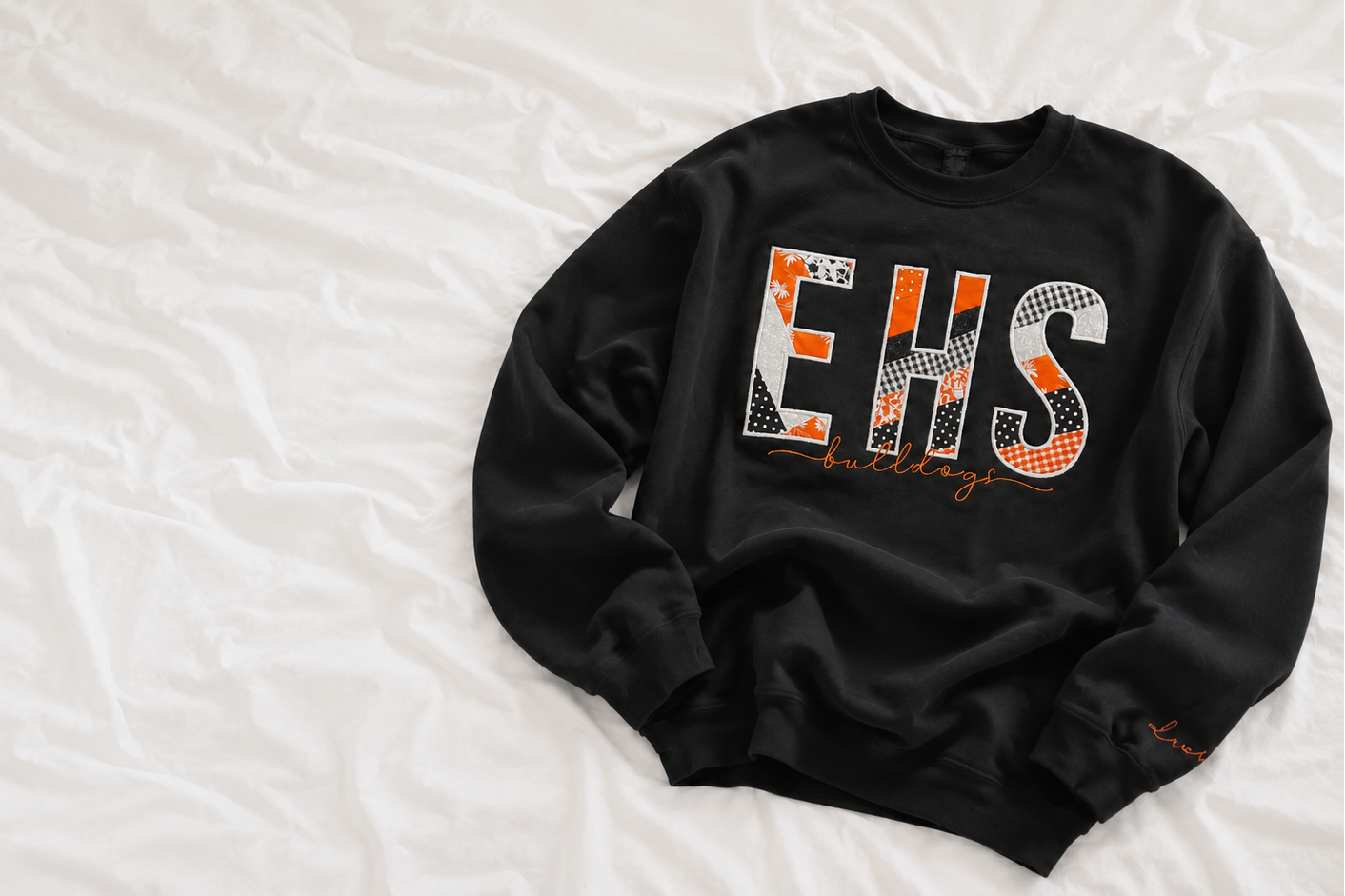 A black crewneck sweatshirt with a large 'EHS' graphic on the chest, laid flat on a white wrinkled fabric background.
