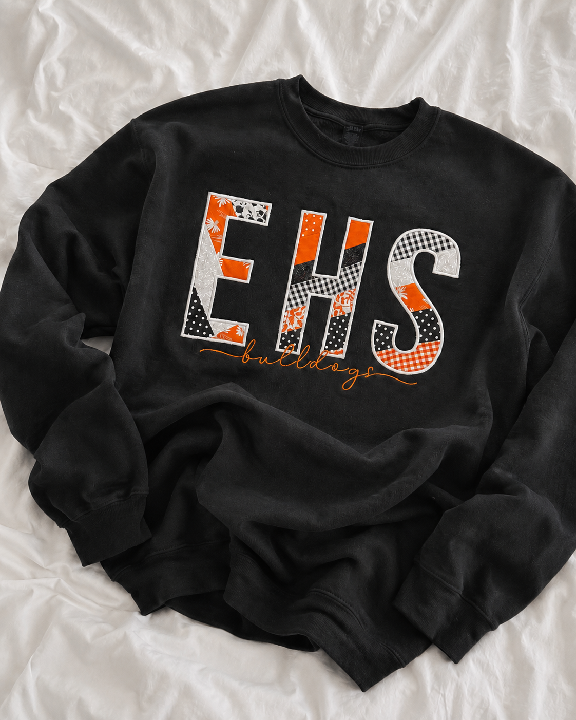 A black crewneck sweatshirt laid flat on a white wrinkled fabric background. The front features a large graphic spelling 'EHS' in a patchwork pattern of orange, black, and white, with 'bulldogs' written in orange script underneath.