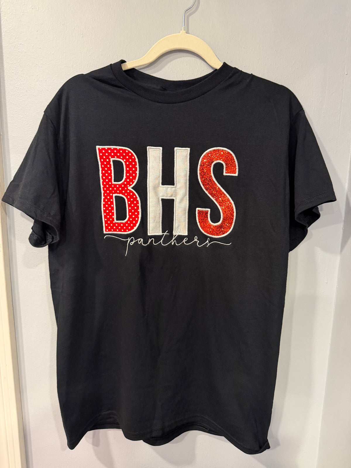 A black short-sleeve t-shirt hanging on a wooden hanger against a light gray wall. The shirt features a large graphic on the front with the letters 'BHS' in red and white, with 'panthers' written in white script underneath.