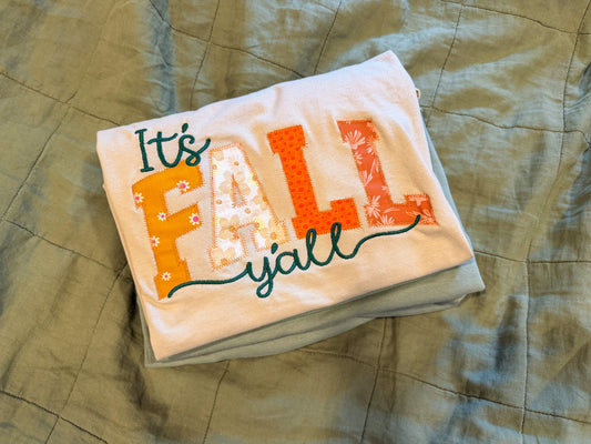 A folded white t-shirt with a colorful 'It's FALL y'all' applique design, resting on a green quilted blanket.