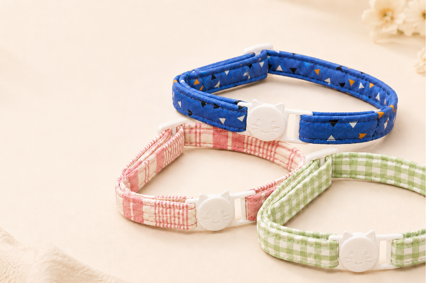 Three adjustable fabric pet collars with white plastic cat-shaped buckles, arranged on a light beige surface. The top collar is blue with small white and orange triangle patterns, the middle collar has a pink gingham pattern, and the bottom-right collar has a light green gingham pattern.