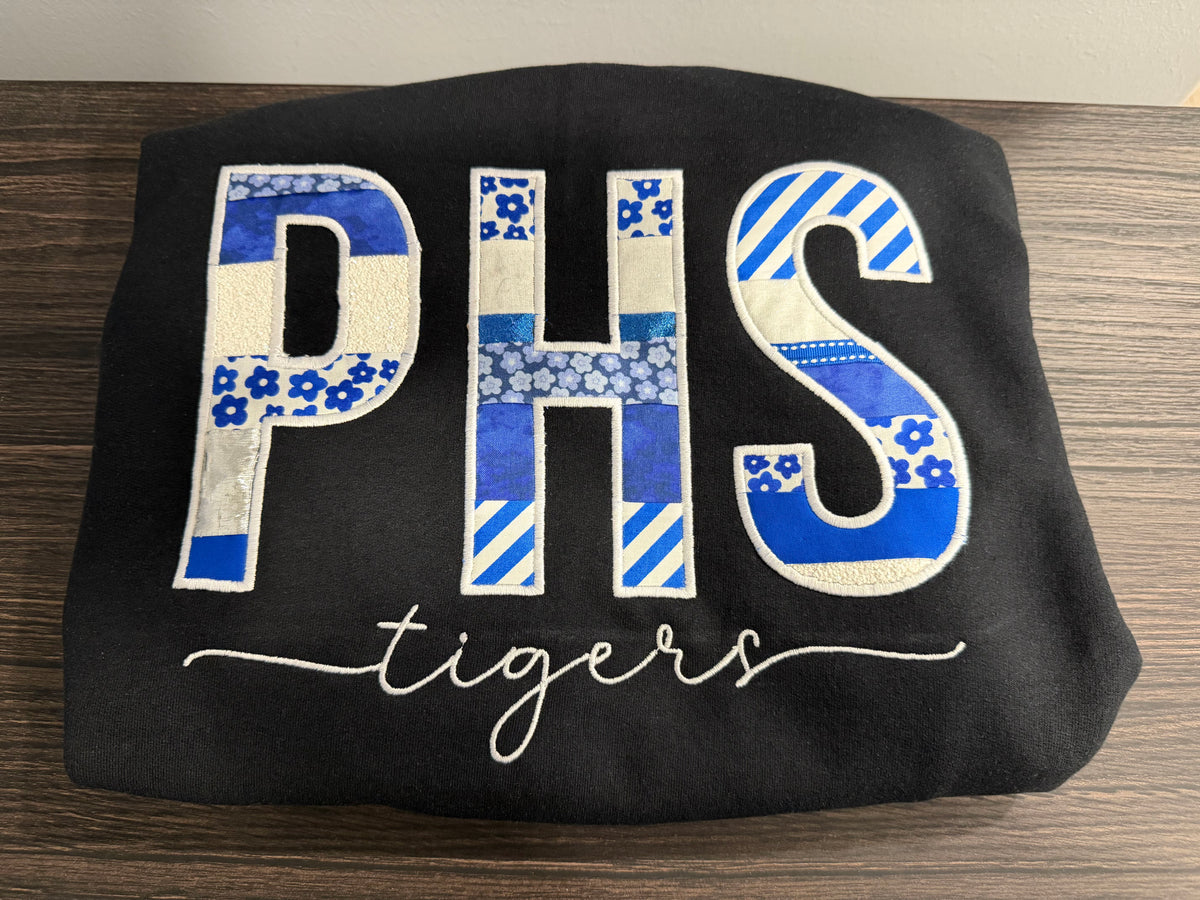 A black sweatshirt laid flat on a wooden surface, featuring large embroidered letters 'PHS' filled with blue and white patterned fabric patches, and the word 'tigers' in white script below.