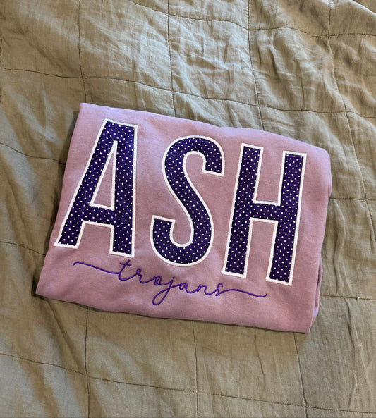 A folded light purple sweatshirt laid flat on a beige quilted blanket. The front features the word 'ASH' in large purple letters with white polka dots and a white outline, with 'trojans' written in purple cursive underneath.