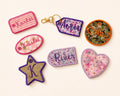 Seven decorative embroidered patches in various shapes (star, oval, rectangular, arch, price tag, heart, circle) with floral and striped patterns, displayed on a light cream background.