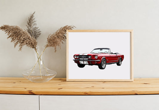 A framed art print of a red classic muscle car convertible with white racing stripes, displayed on a light wood table next to a glass vase with dried pampas grass against a neutral wall.
